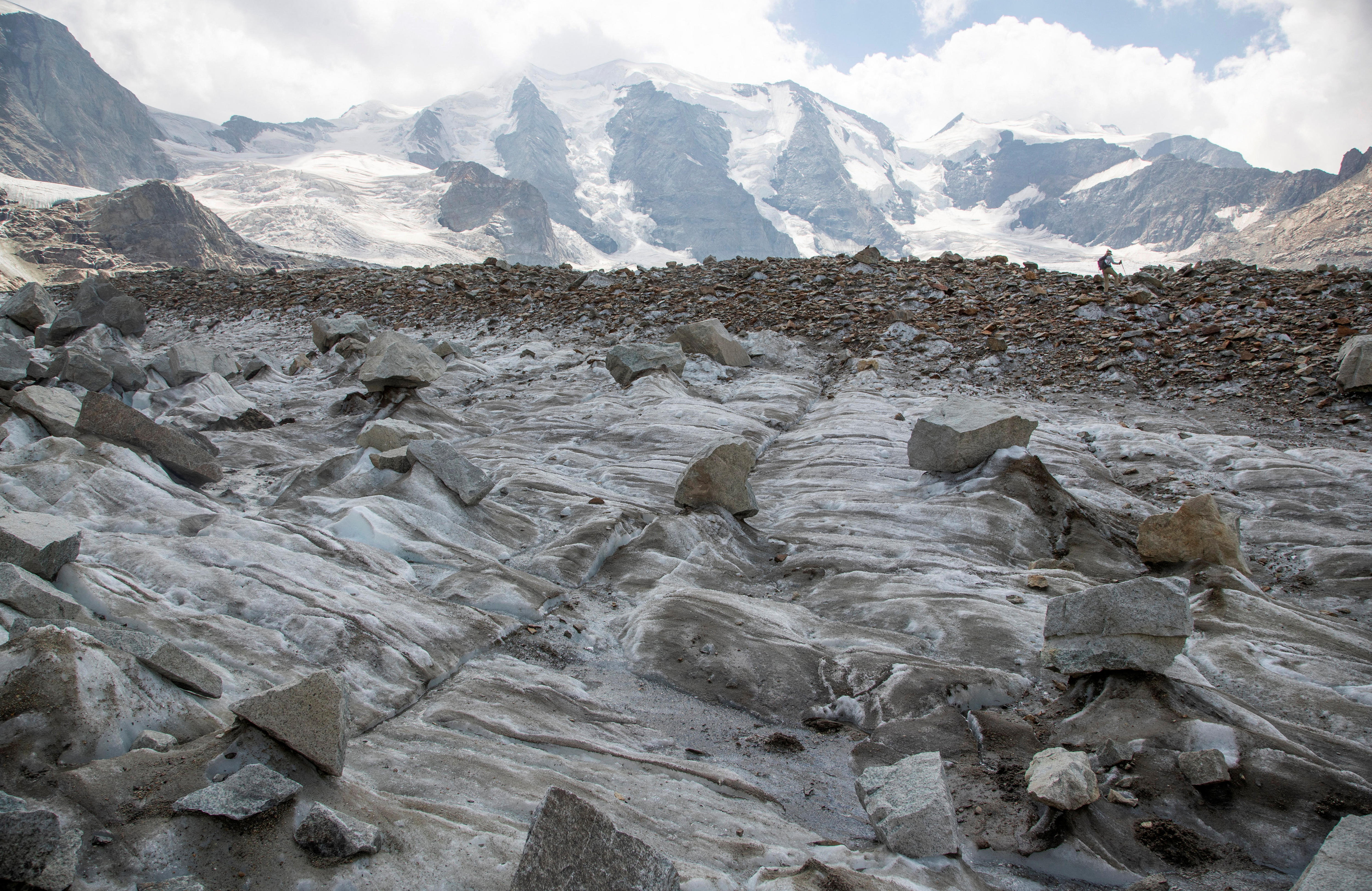 A hiker in the background walks over a glacier. Rocks and ice can be seen in the foreground. 