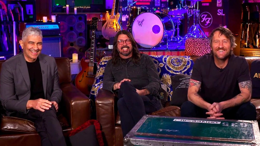 Pat Smear, Dave Grohl and Chris Shiflett of Foo Fighters sit on a couch in front of a drumkit and amps