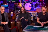 Pat Smear, Dave Grohl and Chris Shiflett of Foo Fighters sit on a couch in front of a drumkit and amps