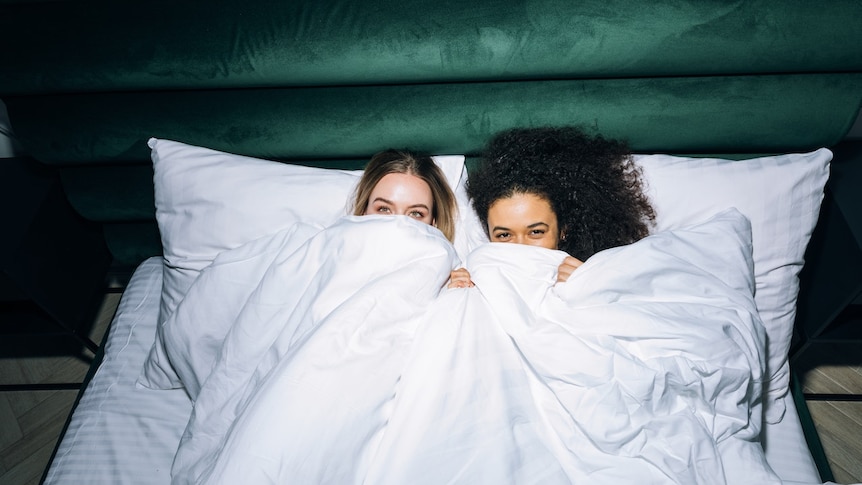 Two young women lying in a bed with covers pulled up to their eyes.