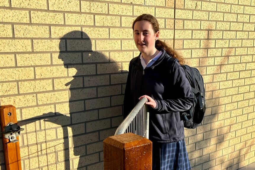 A teenage girl, wearing a backpack and school uniform, opens a gate beside a brick wall.