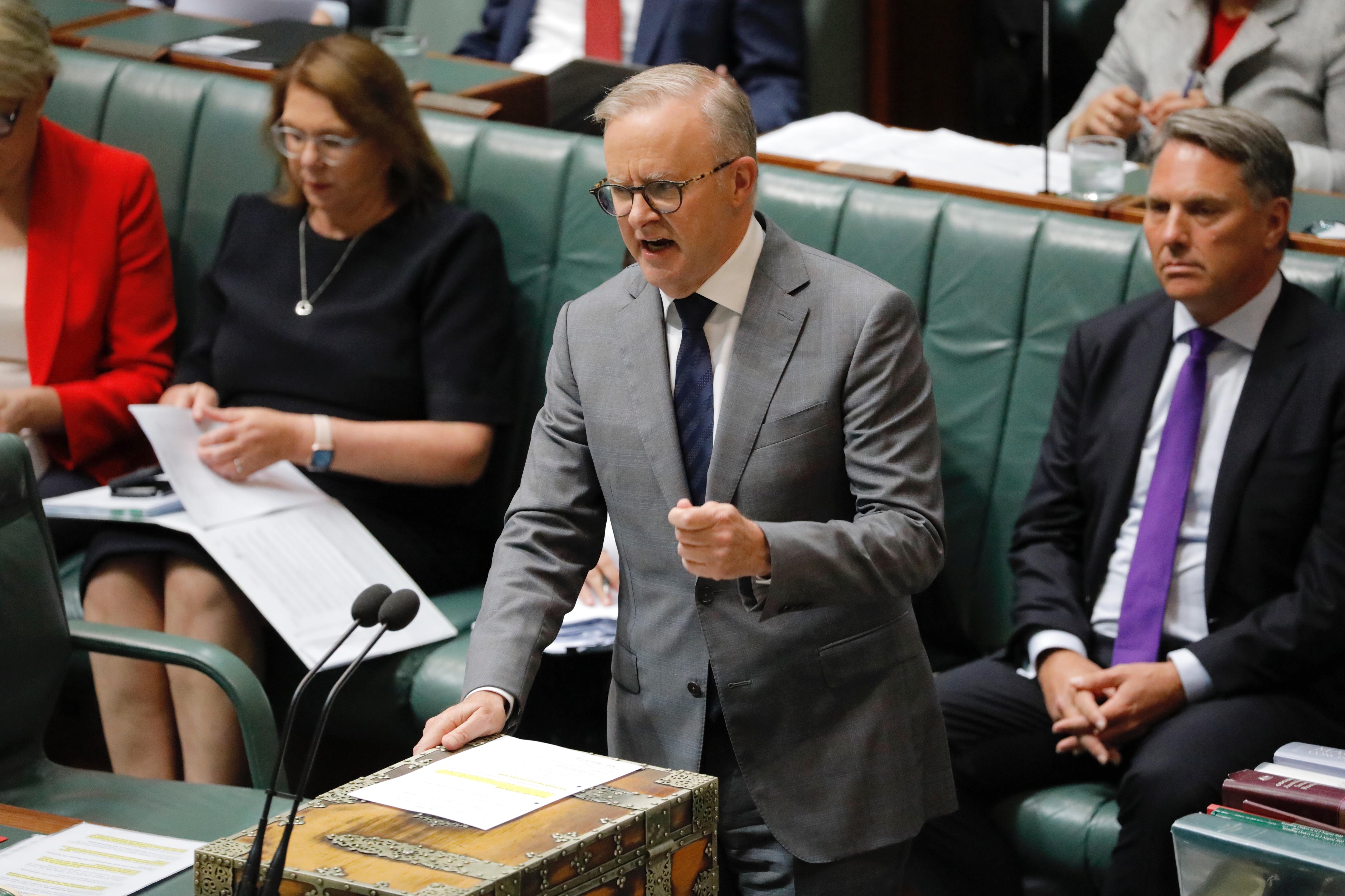 A man in a suit and tie stands and speaks in parliament, with government members sitting behind him