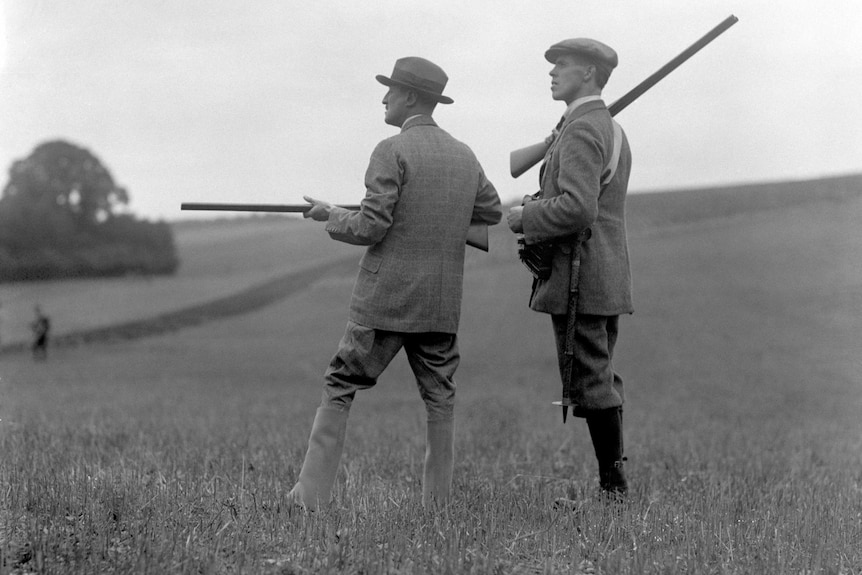 Black and white image of two men in early 20th century English hunting garb, holding rifles, staring off into the distance.