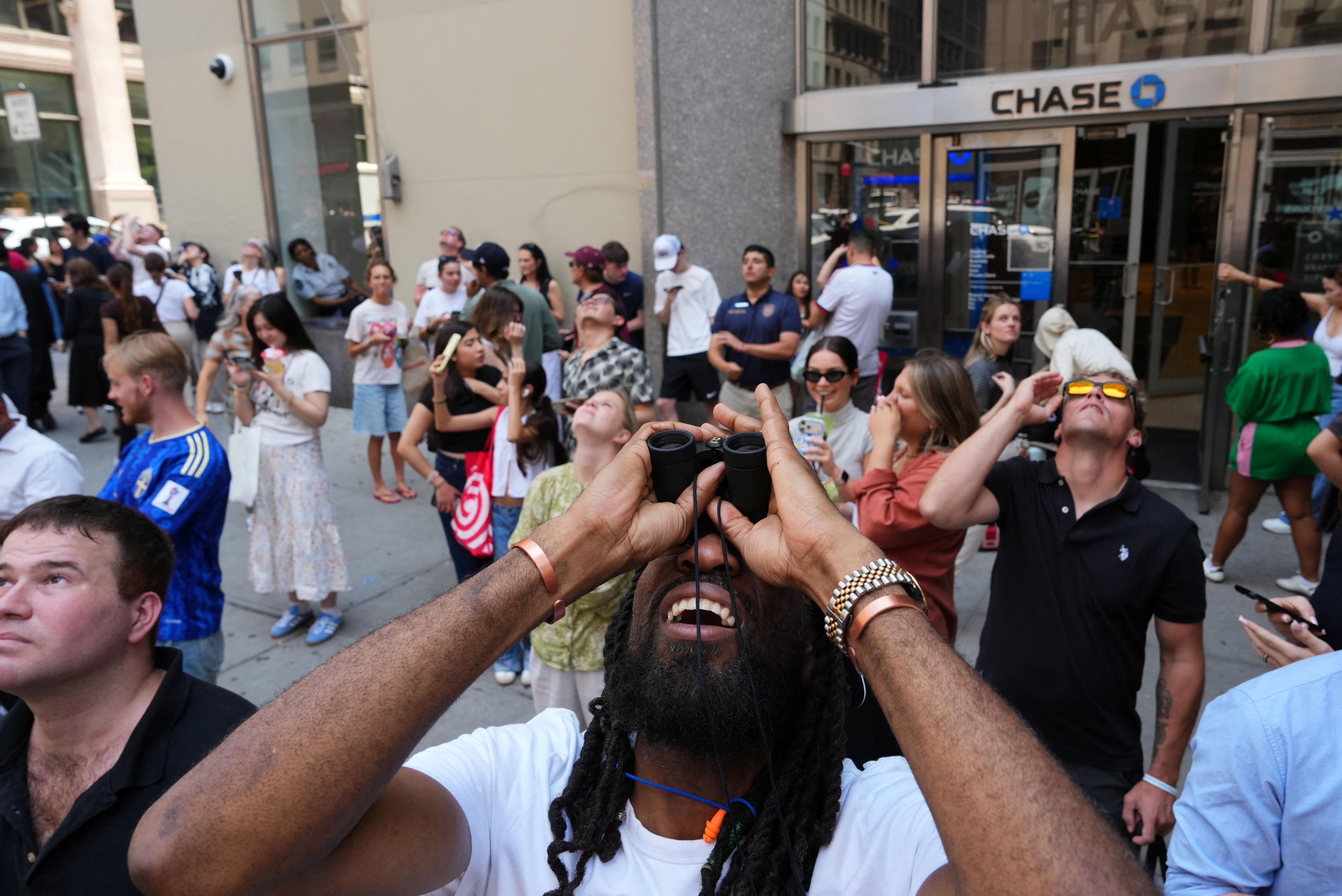 Tourists and bystanders looking upward, some with binoculars, on a footpath outside grey tower buildings.
