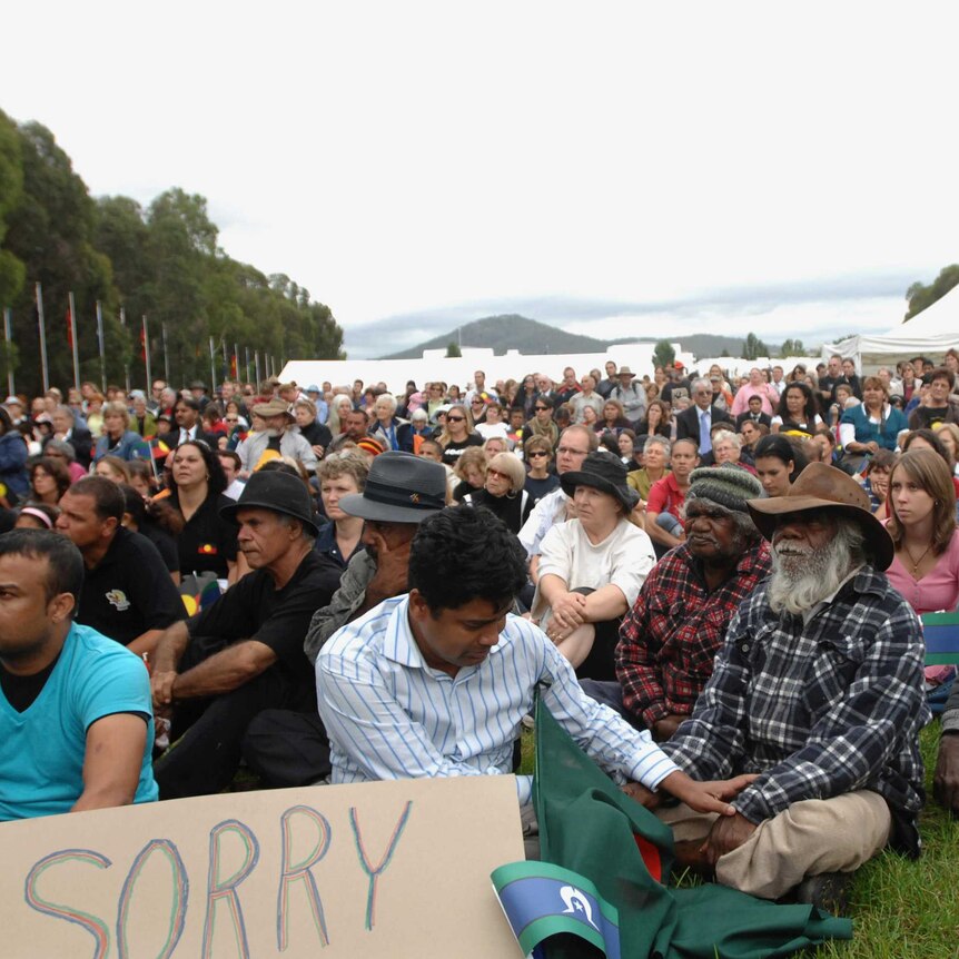 Thousands sit on the grass outside Parliament House in Canberra, including many First Nations people. The mood is sombre.