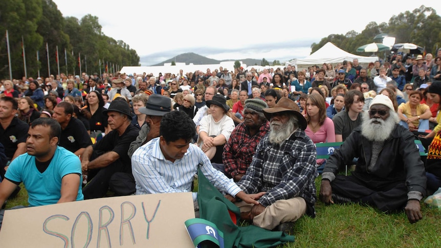 Thousands sit on the grass outside Parliament House in Canberra, including many First Nations people. The mood is sombre.