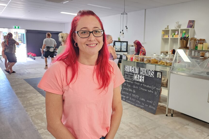 A young woman in a pink shirt stands inside a cafe.