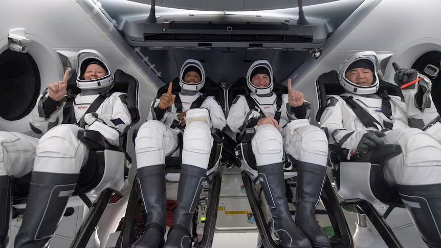 Four astronauts strapped into seats in a capsule all holding up one finger.