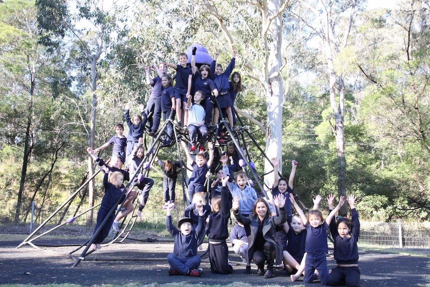 Around 20 students sitting on a playground spider equipment cheering at the camera, with principal Erin Eade.
