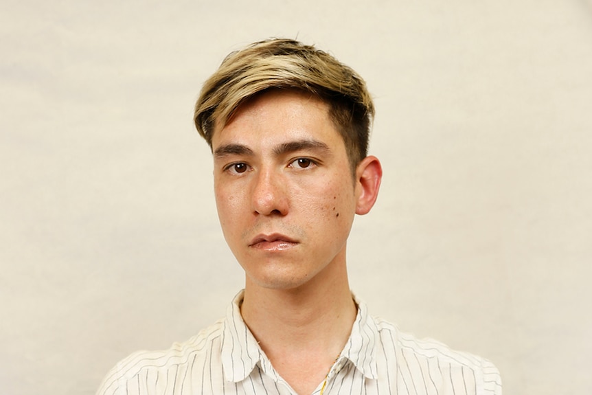 Thai-Australian artist Nathan Beard stands with serious expression in front of wall in oatmeal pinstriped button up shirt.