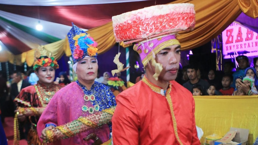 A procession of bissus walk past the camera in bright and colourful clothing.