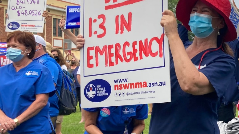 Two women wearing blue scrubs, one holding a white sign with red letters on it