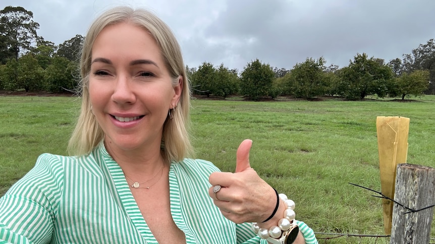 A young woman with blonde hair smiles and gives a thumbs up with green grass behind her.