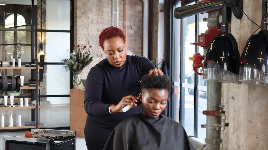 Rumbie stands in her salon, styling her customer's hair. Rumbie and her client have short afro hairstyles. Rumbie's is dyed red.