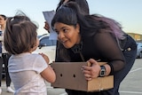 Young woman in black bends down to chat to a toddler while holding a box with holes in a car park. People behind.