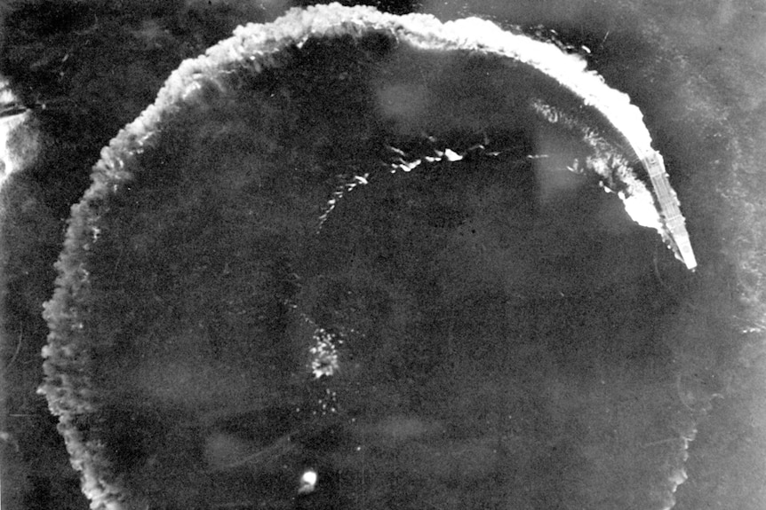 A black and white aerial photo of a warship turning in a circle at sea