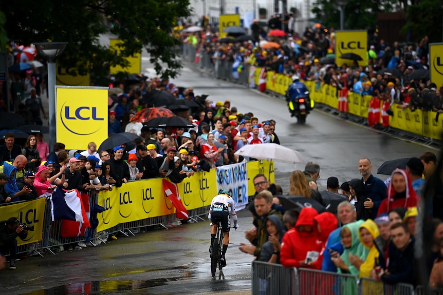 A bike rider races along a wet road while fans look on behind barricades
