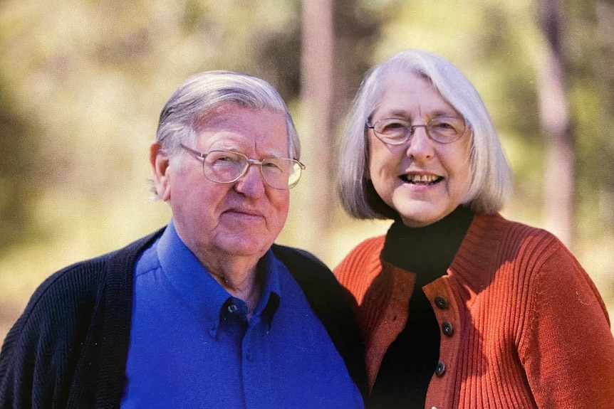 An elderly couple stand together in blue and red jumpers.
