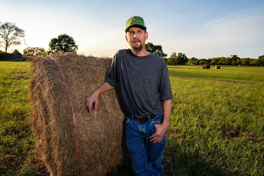 A man in a baseball cap leans against a hay bale