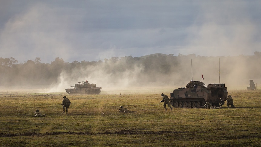 Dust rises over a field as soldiers and a tank move across it.