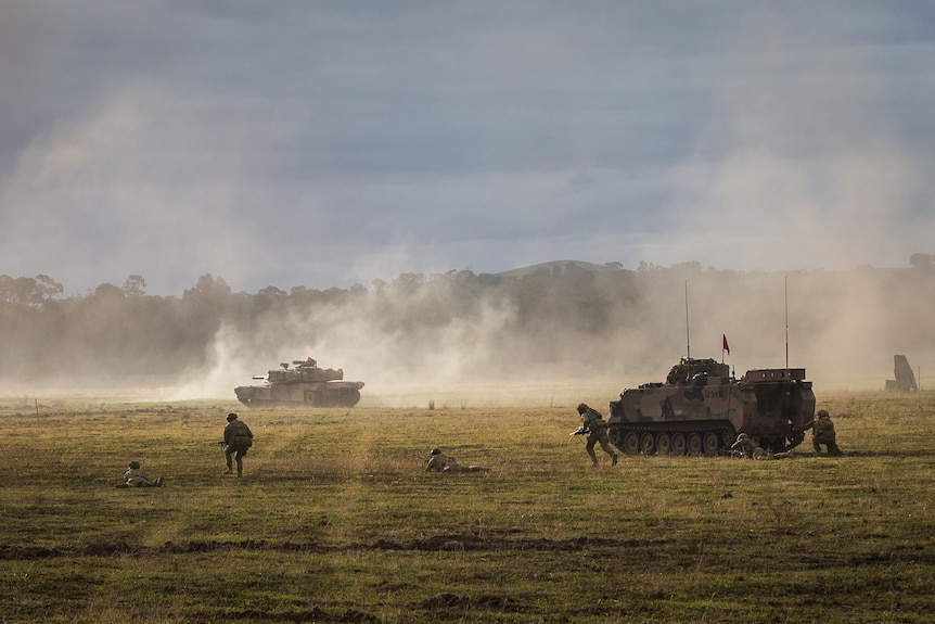 Dust rises over a field as soldiers and a tank move across it.