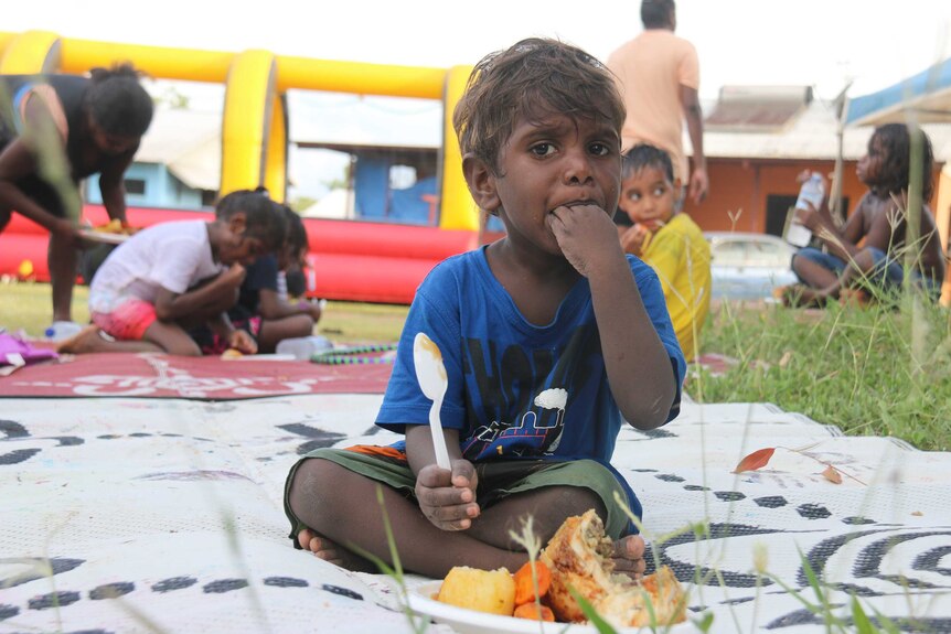 A little boy sitting on a mat on the grass shovels some roast chicken dinner into his mouth.