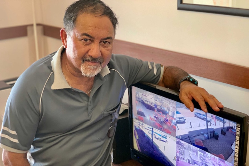 A man leans on a television screen that shows a four-way split screen of CCTV footage.