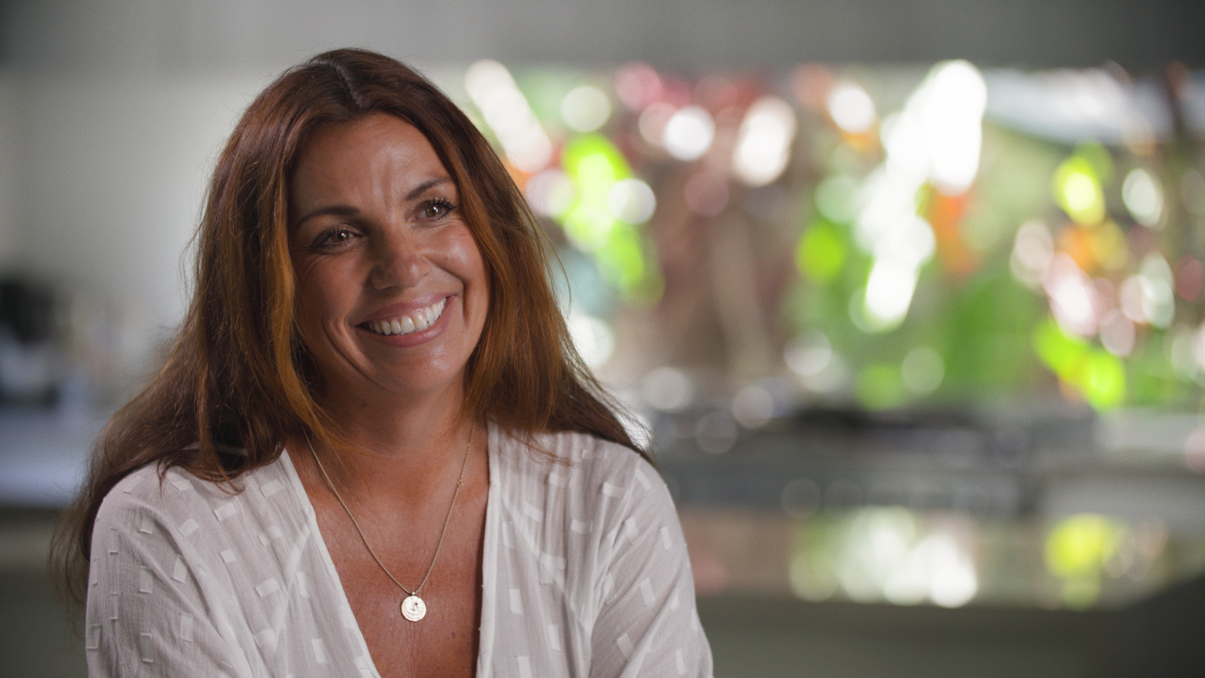 A woman with long brown hair wearing a white top sits in a kitchen and smiles.