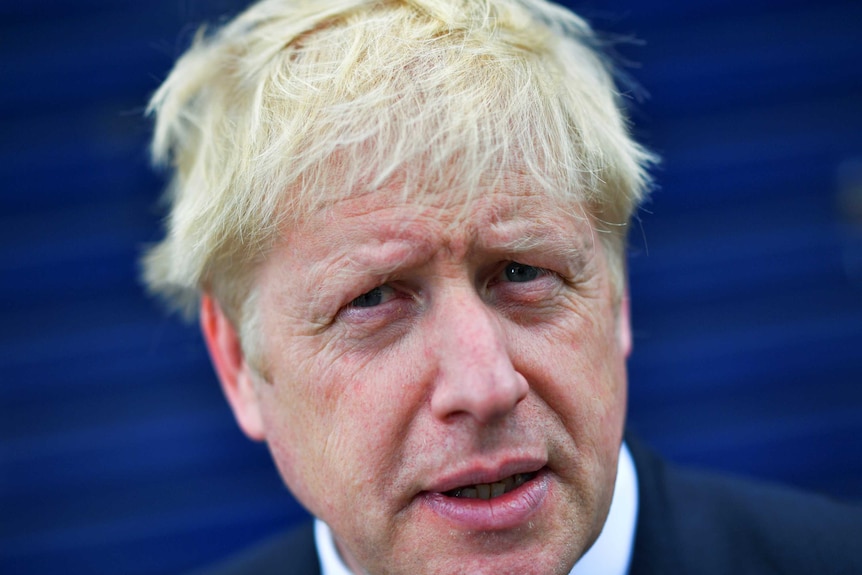 A blond man in a suit holds an ice cream with a tiny British flag stuck in it