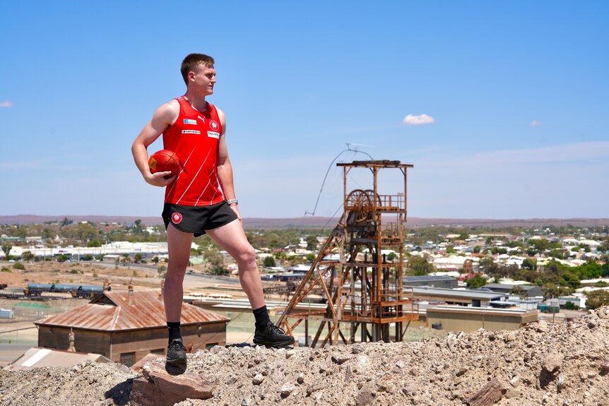 A young man wearing a red singlet, black shorts holds a football while standing on top of Broken Hill Line of Lode. 