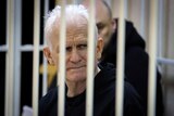 A thin-looking older white man with white hair sits in a barred cell wearing black clothing.