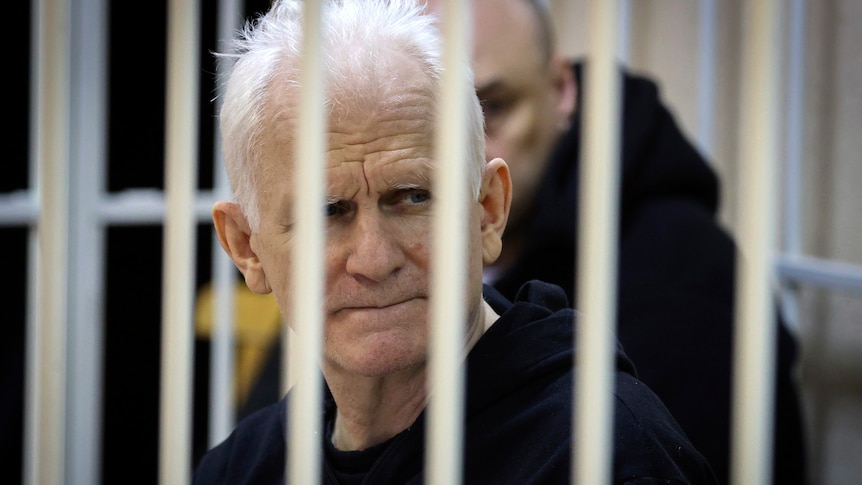 A thin-looking older white man with white hair sits in a barred cell wearing black clothing.