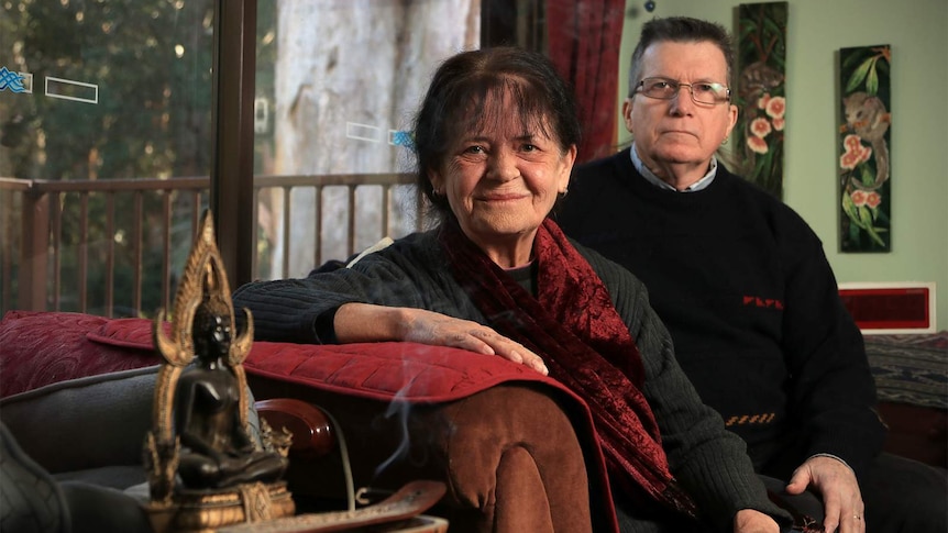 A woman with dark hair sits with a man wearing glasses. They're on a red lounge and the woman is smiling.