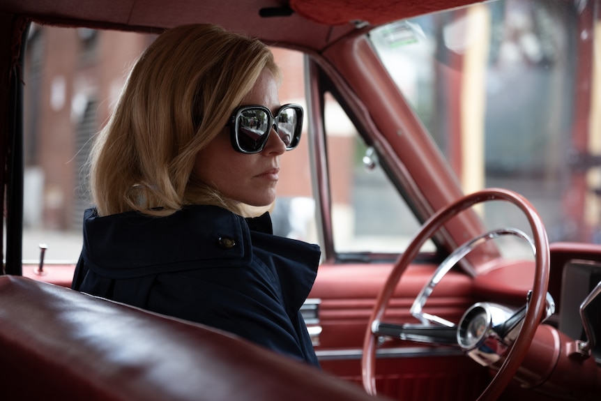 A blonde middle-aged woman is sitting in her 60s-style car. She's wearing large, dark sunglasses and looks determined.