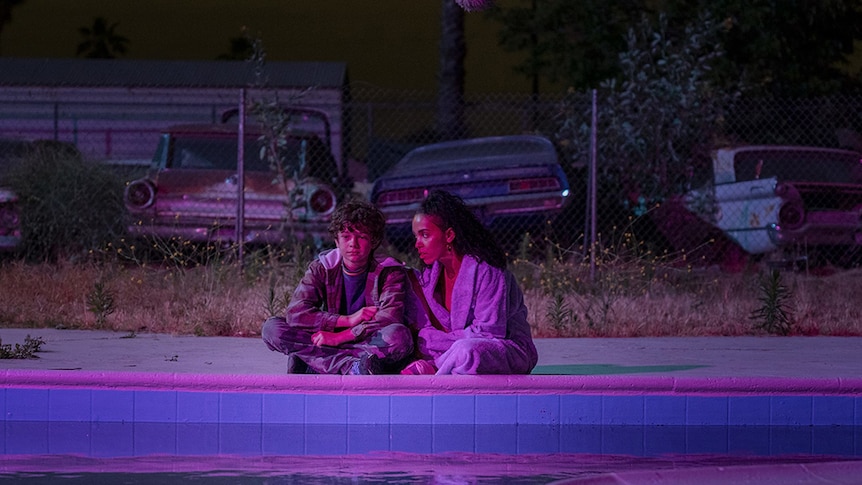 A teenage boy in jeans and zip up hoody sits at edge of pool with woman in bathrobe, behind them a wire fence and vintage cars.