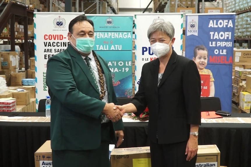 A man and a woman in formal clothes and face masks shake hands as they stand inside a full warehouse.