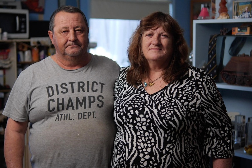 A man and a woman stand inside their house looking at the camera with neutral expressions.