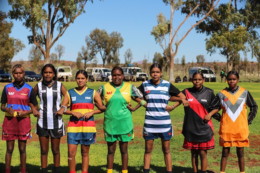 A row of young Aboriginal women line up arm in arm wearing colourful football jumpers on a football ground