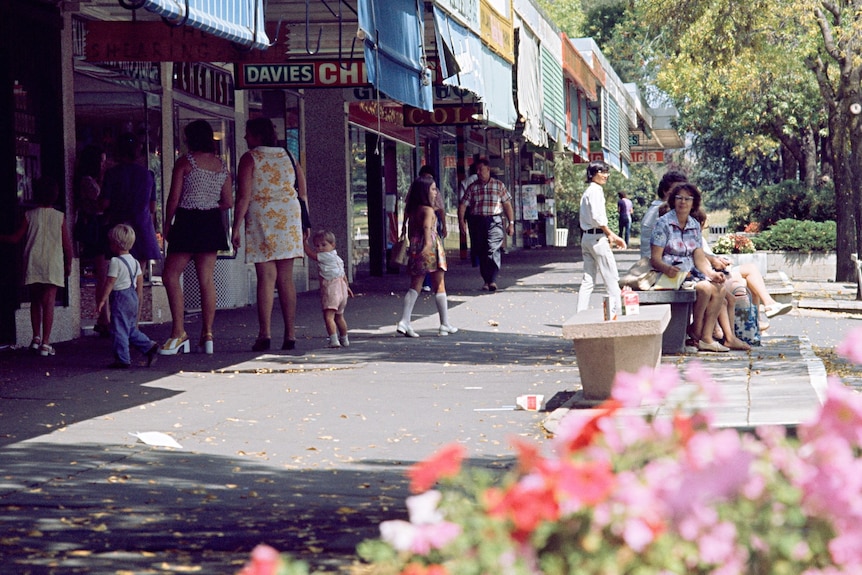 an old photo of a woman sitting on a bench in a busy street in kingston