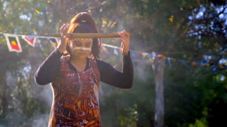 An Aboriginal woman with paint on her face performing a dance holding a stick over her head