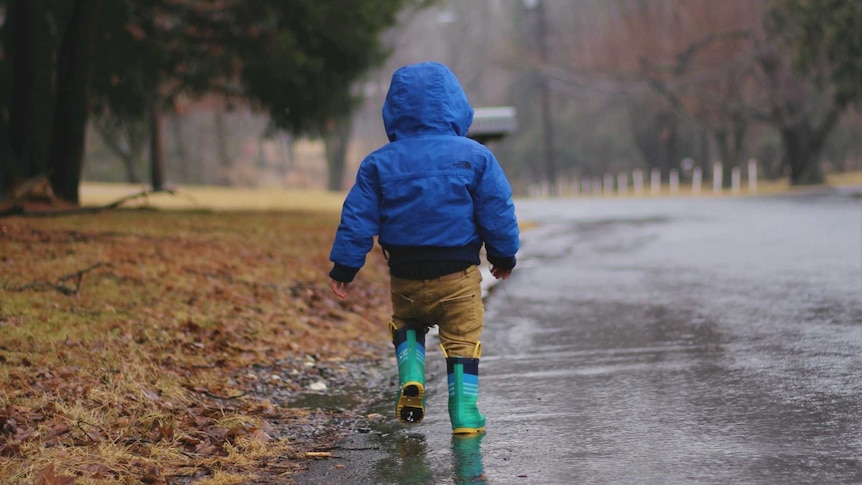 A toddler walks through the rain wearing a jacket and gumboots