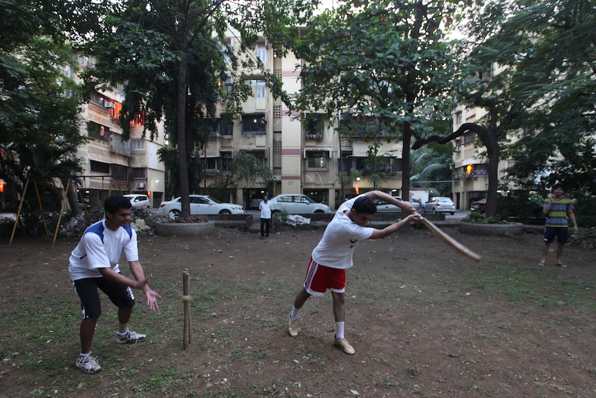 A game of cricket is played on an area of grass next to a street in India with an apartment complex in the background.