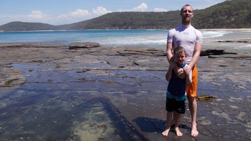 A man in rash-shirt with his arms around child at rock pool with beach in the background.