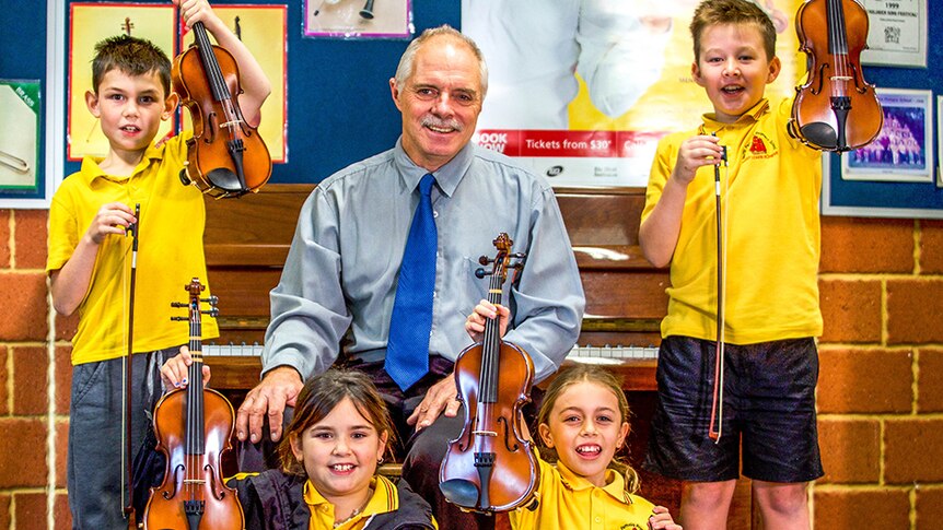 Schoolchildren hold up violins while a teacher sits in front of a piano in a primary school music room.