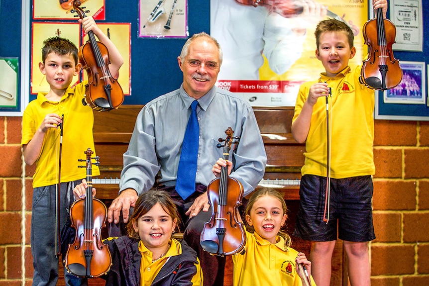 Schoolchildren hold up violins while a teacher sits in front of a piano in a primary school music room.