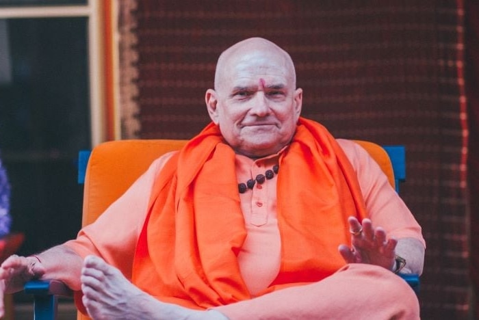 Russell Kruckman looks into the camera as he sits on a chair, dressed in orange clothing.