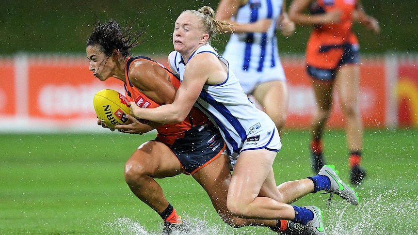 An AFLW player grimaces as she is tackled from behind, as she splashes through a sodden ground.
