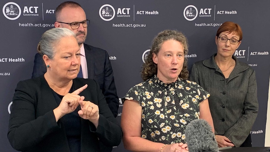 A woman in a floral shirt stands in front of a microphone at a press conference.