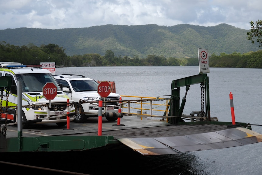 An ambulance and four wheel drive move across the Daintree river on a ferry, with mountains in the background.