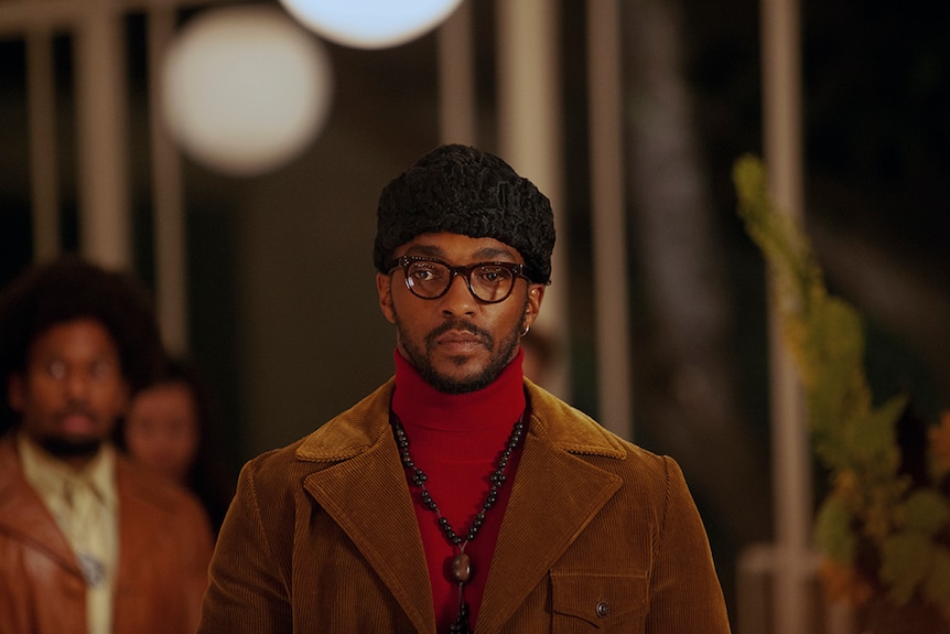 A standing man with neutral expression wears black hat, square framed glasses, red turtleneck and caramel corduroy jacket.